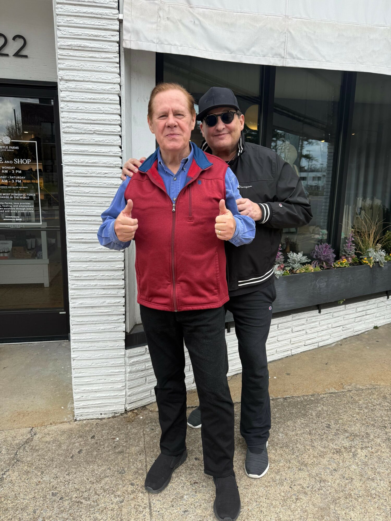 Two men standing outside, one in a black jacket and sunglasses, the other in a red sweater and blue shirt, both giving thumbs up.