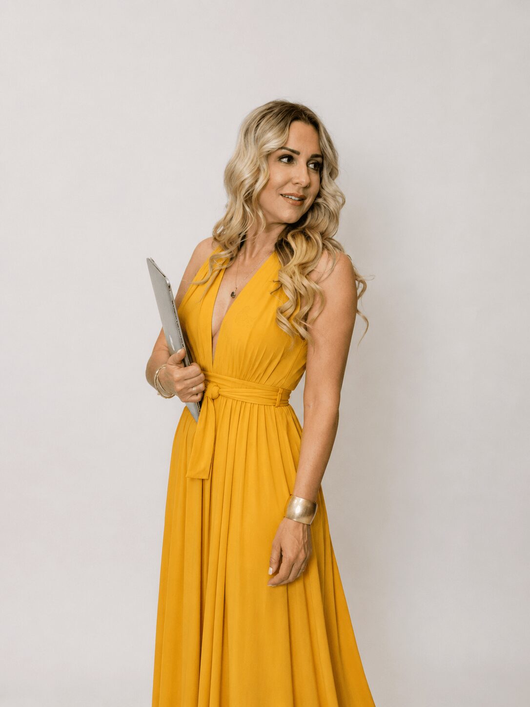 Woman with long blonde hair in a yellow dress holding a tablet, standing against a plain background.