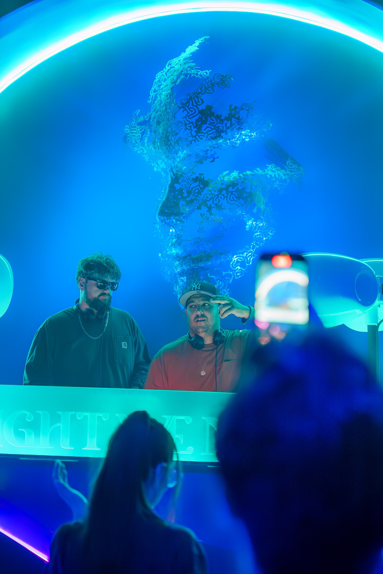 Two people stand behind a glass display with a large globe and water splash projection, with a person taking a photo in foreground.