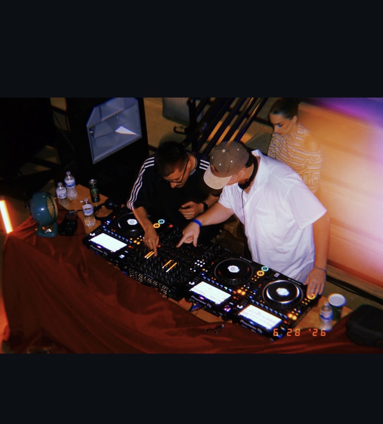 Two people are DJing at a table with equipment, bottles, and cups, while a woman watches nearby. Colorful lighting is present.