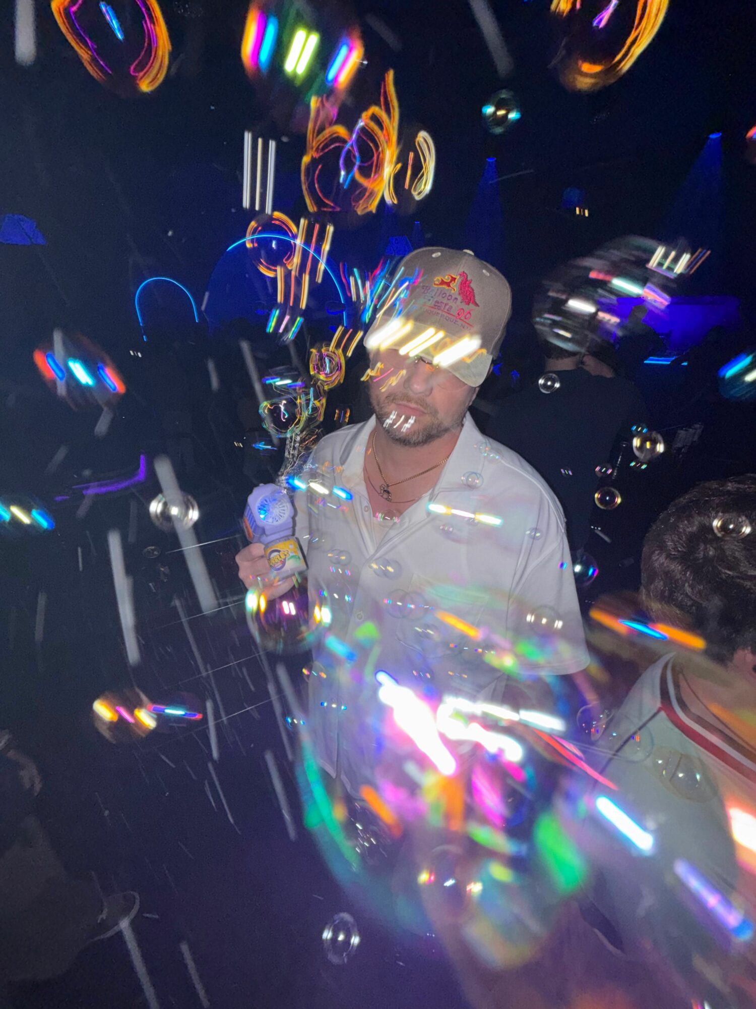 Person wearing a white cap and shirt, surrounded by colorful light reflections and bubbles, in a dark environment.