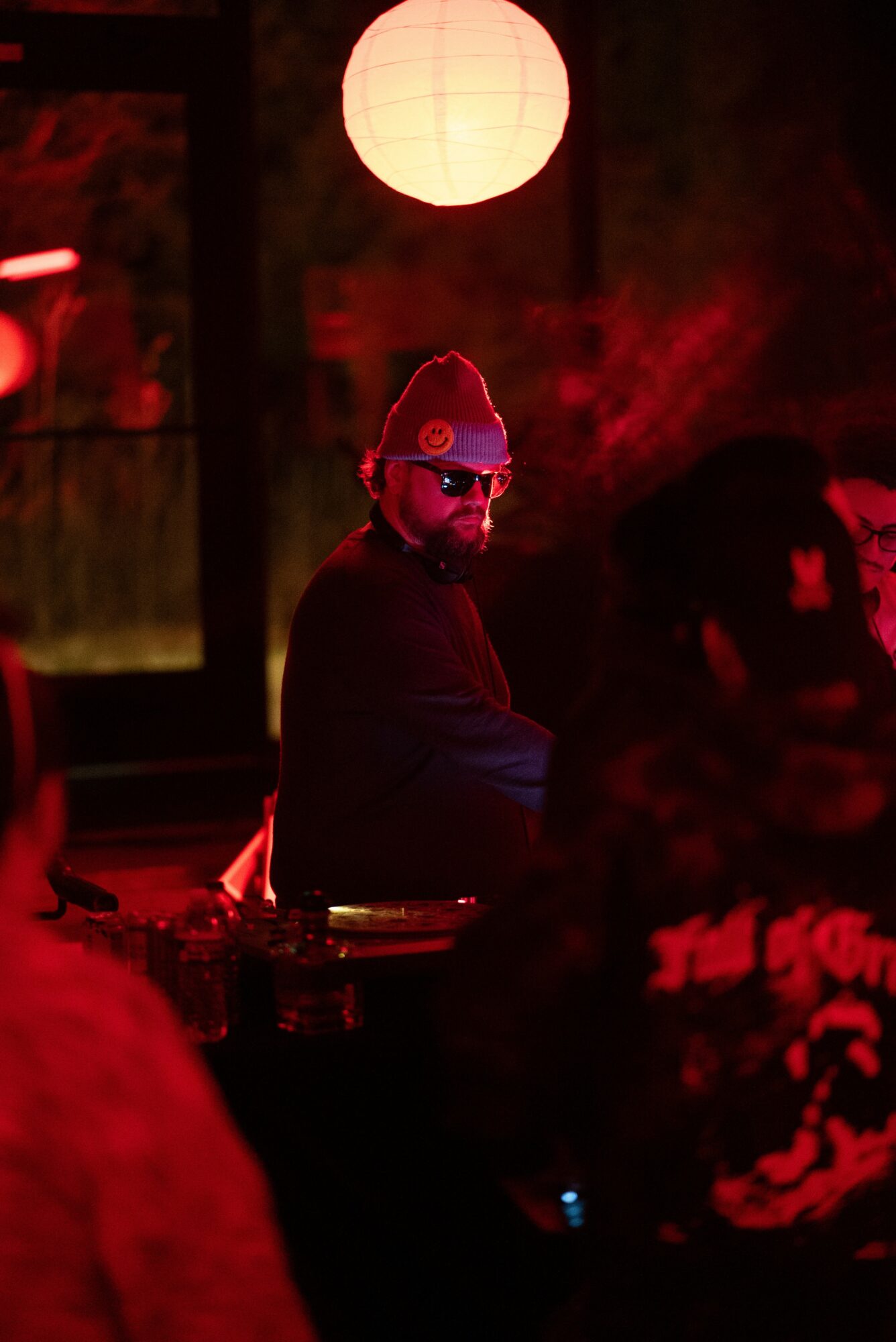 Person wearing sunglasses and a beanie in a dimly lit space with red lighting and a paper lantern overhead.
