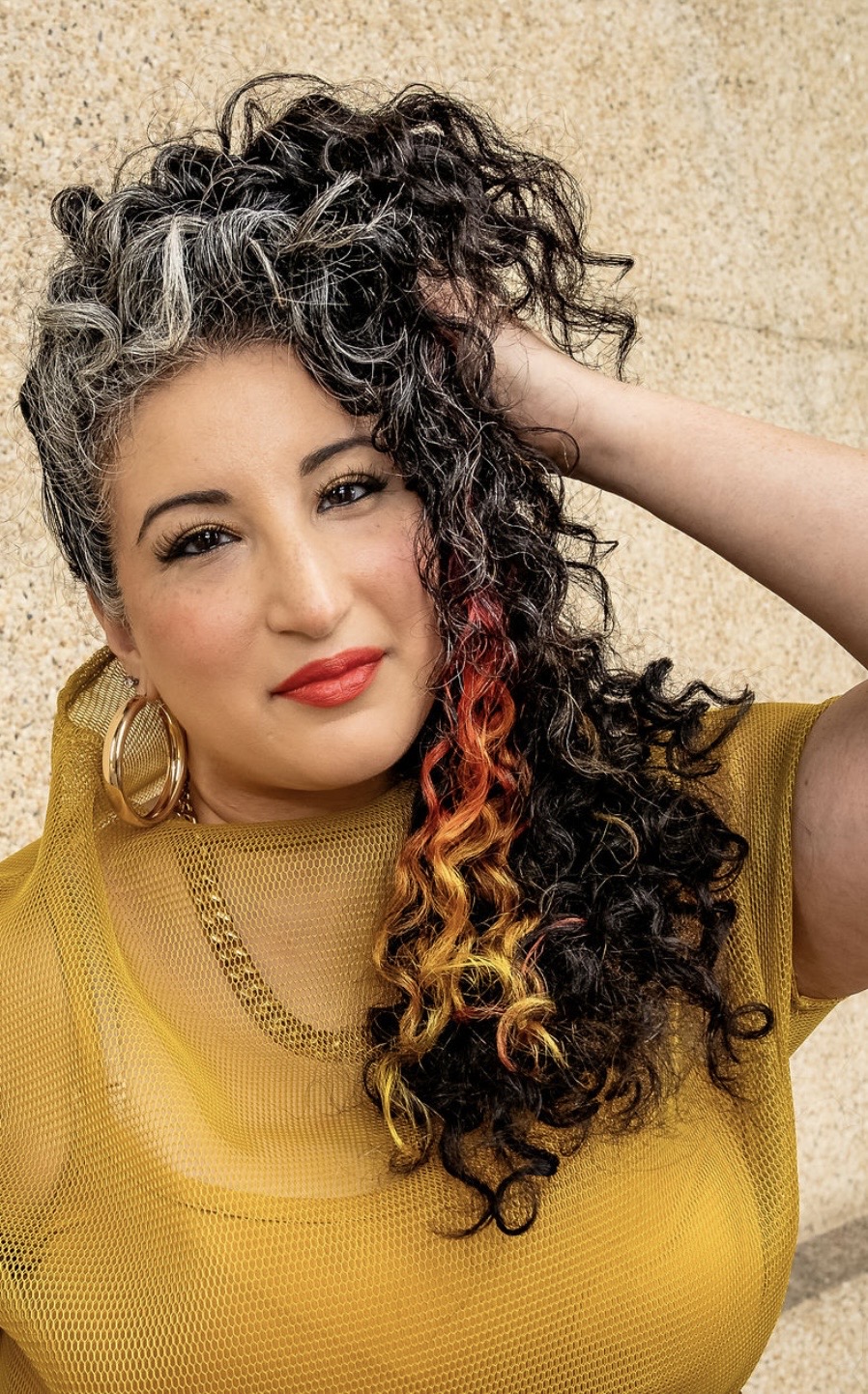 Woman with curly hair, wearing a yellow top and gold jewelry, posing with hand behind head, smiling.