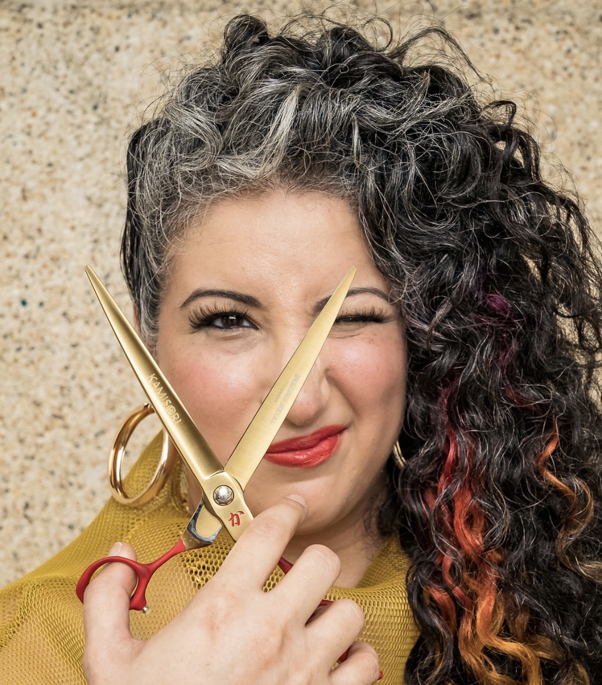 Woman with curly hair, one side gray, the other dark with red tips, holding scissors near her face, winking, smiling, against a textured background.