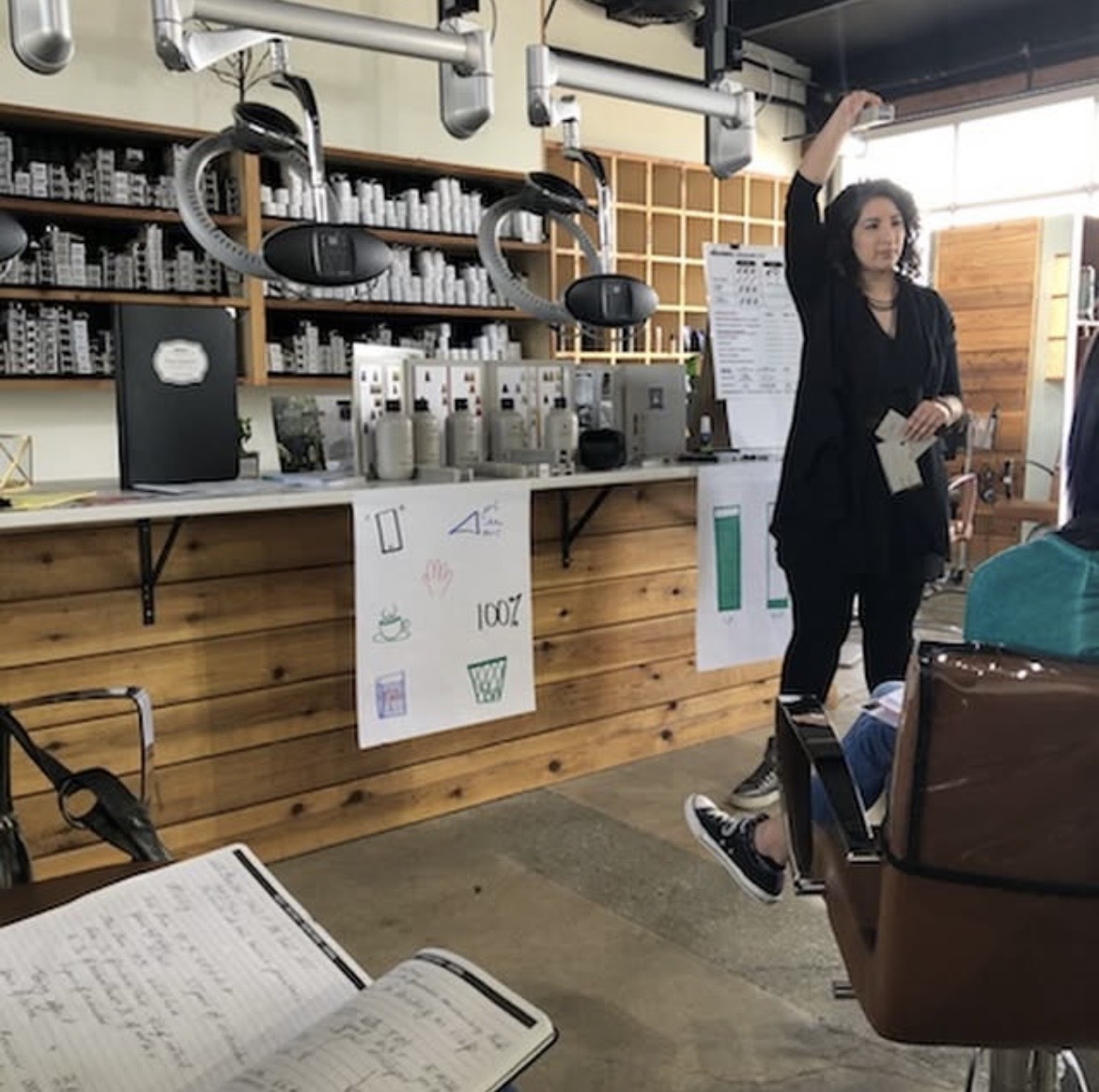 Woman standing and gesturing in a room with shelves, equipment, and posters, holding a notebook. Audience seated nearby.