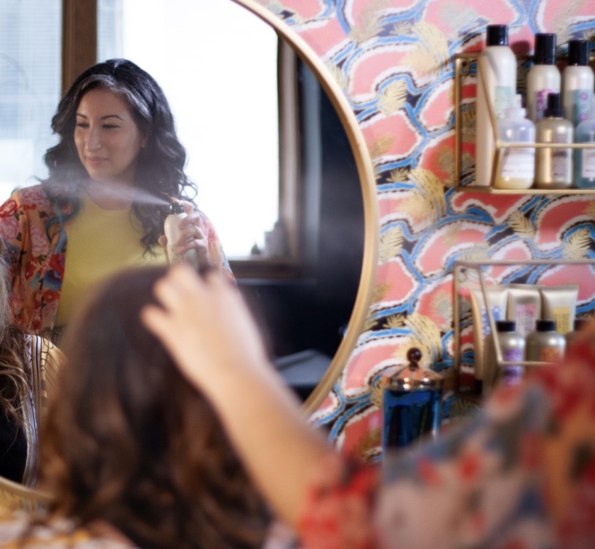 Woman looking at her reflection in a mirror, with colorful wallpaper and shelves of bottles behind her.