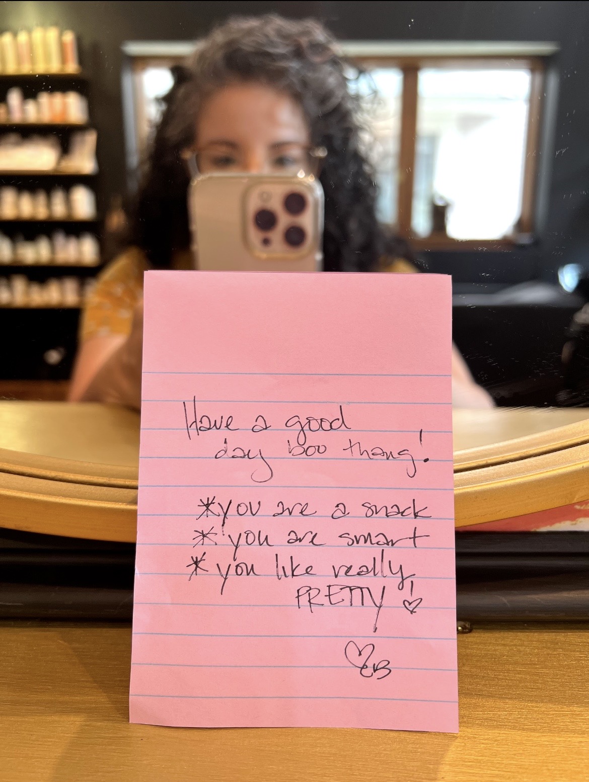 Person with curly hair taking a mirror selfie in a room with shelves, holding a pink note with handwritten message, partially obscured by a phone.