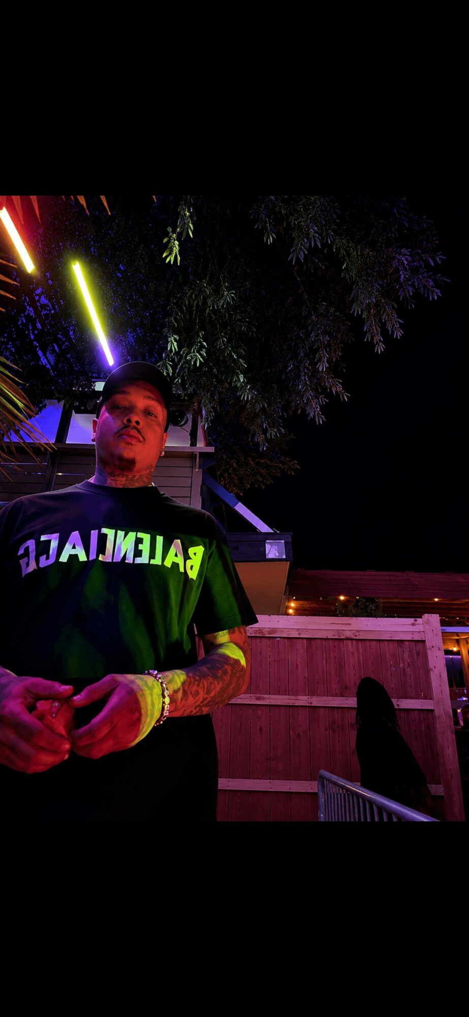 Person standing outdoors at night with colorful neon lights and trees in background.