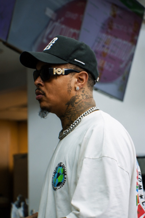 Man with tattoos, sunglasses, black cap, and white shirt, standing indoors with a screen in the background.