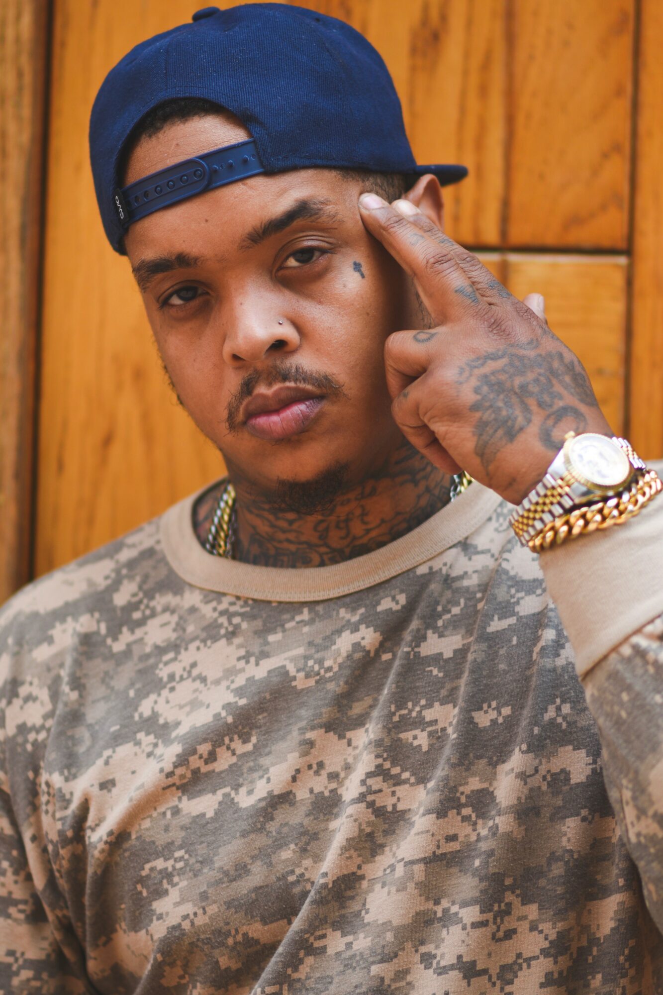 Young man with a blue cap, tattoos, and jewelry, pointing to his face, standing in front of a wooden background.