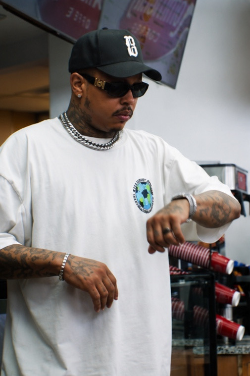 Man wearing a black cap, sunglasses, white t-shirt, and jewelry, looking at his watch in a store.