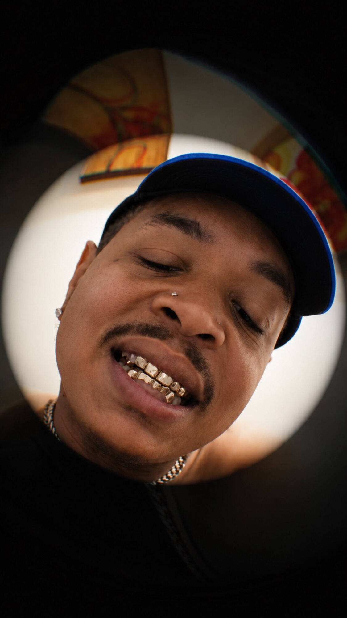 Close-up of a person wearing a cap, showing teeth with a gold grill, smiling slightly, with a dark background.