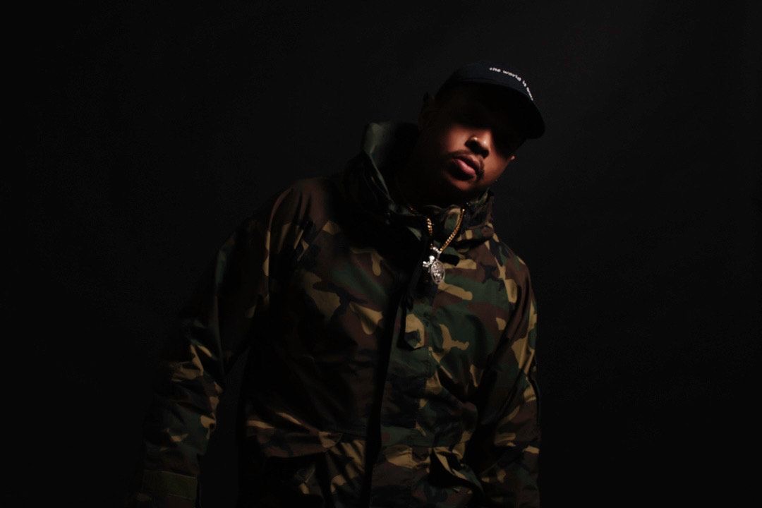 Person wearing a camouflage jacket and cap, facing slightly to the right, against a dark background.