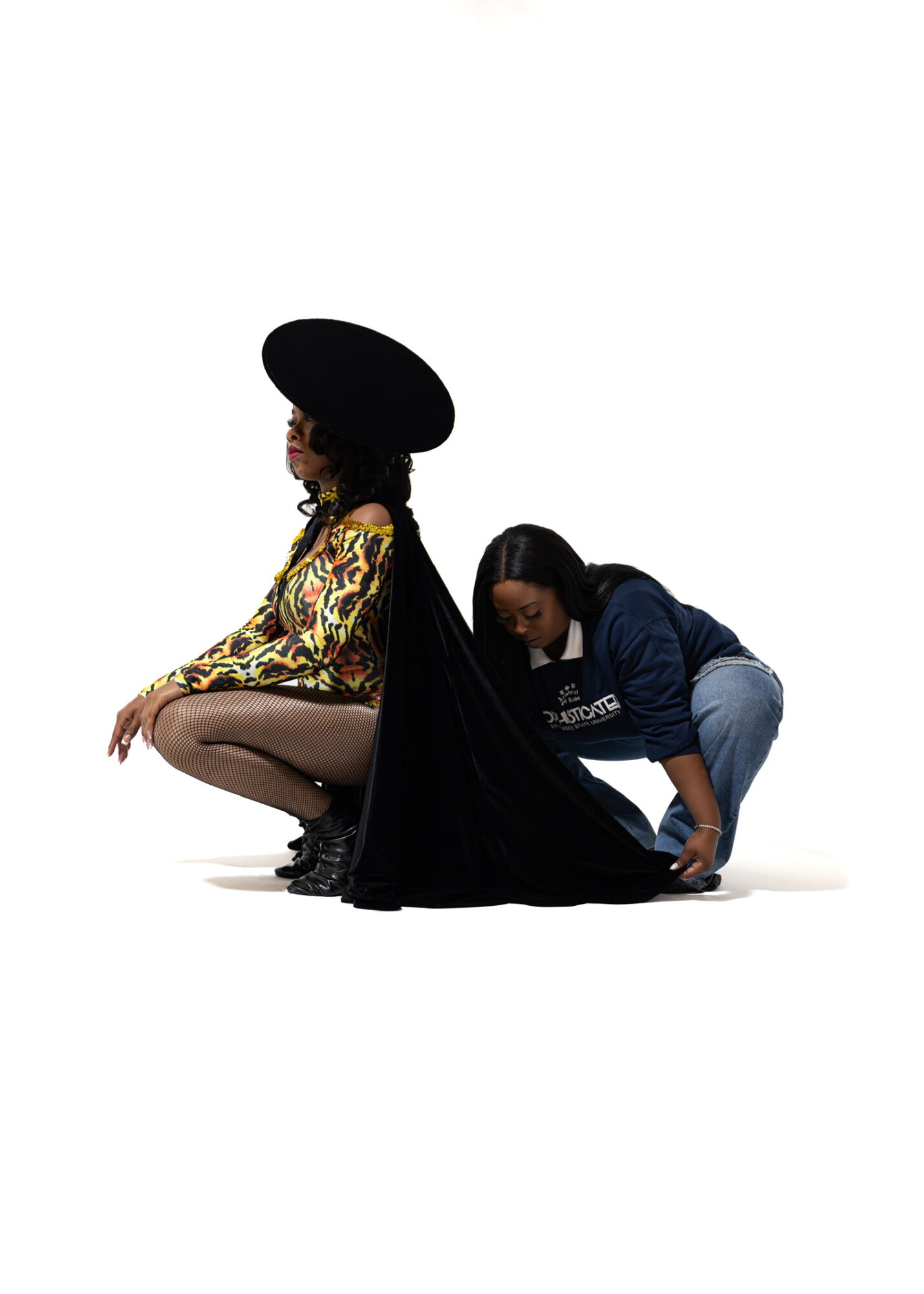 Two women, one sitting with a large hat and patterned dress, the other crouching behind her, adjusting her shoe.