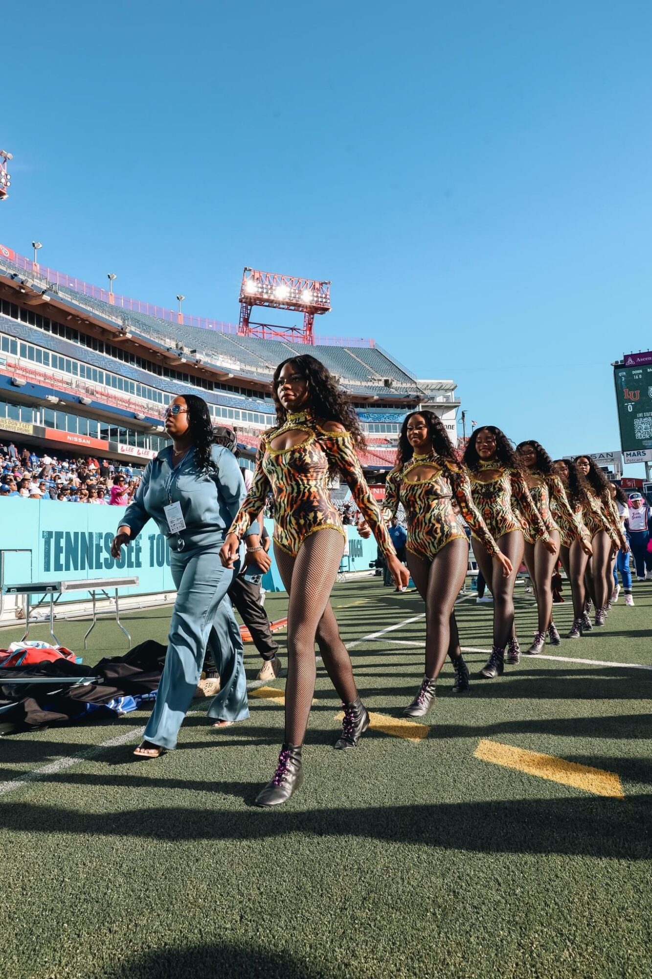 Group of cheerleaders in matching outfits performing on a football field with a stadium and blue sky in background.