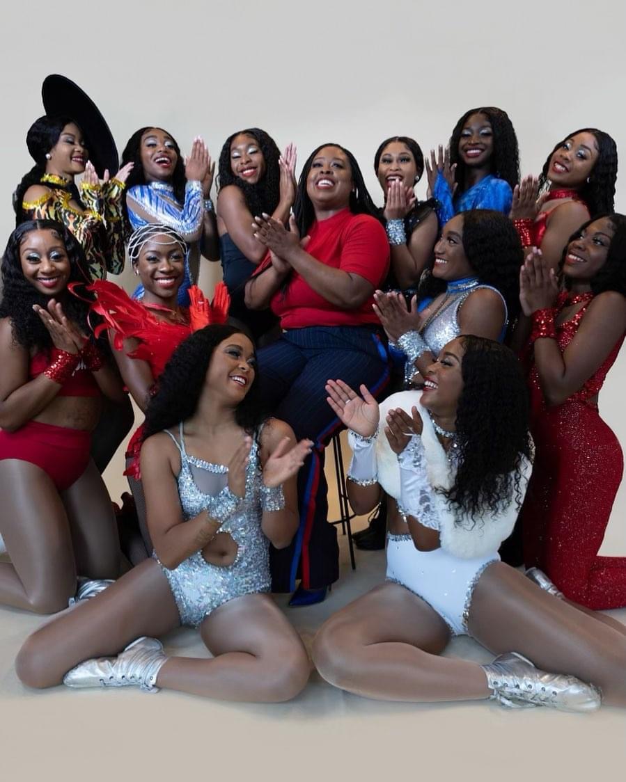 Group of women in colorful costumes posing together, some sitting and some standing, smiling and clapping, against a plain background.