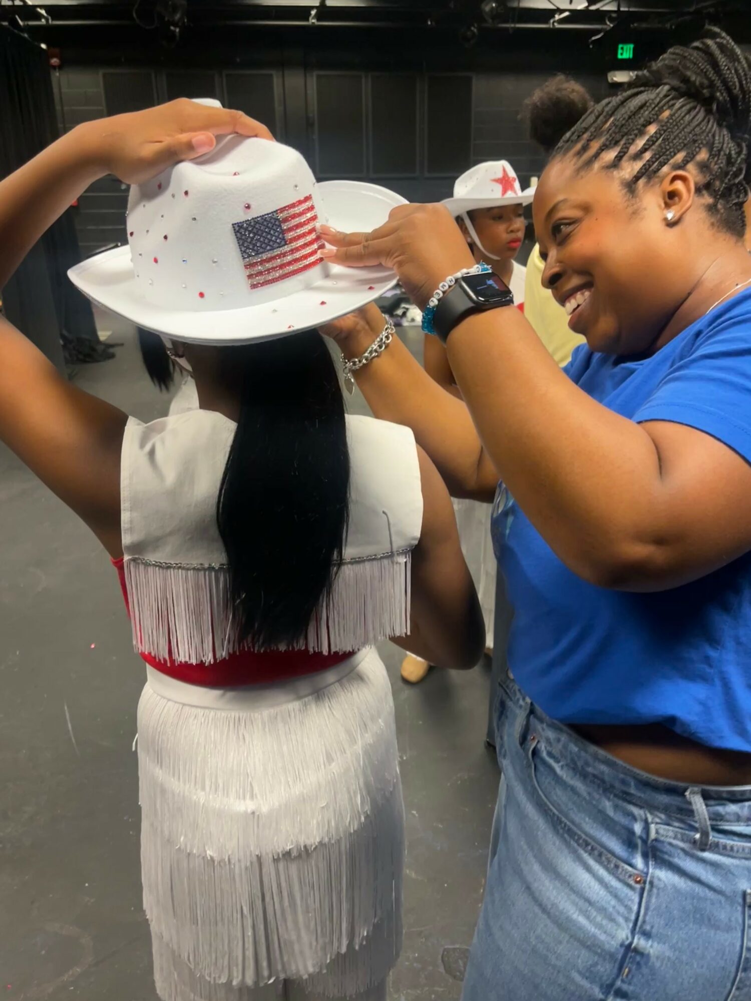 Two women smiling and wearing hats, one with a white hat with an American flag, in an indoor setting.