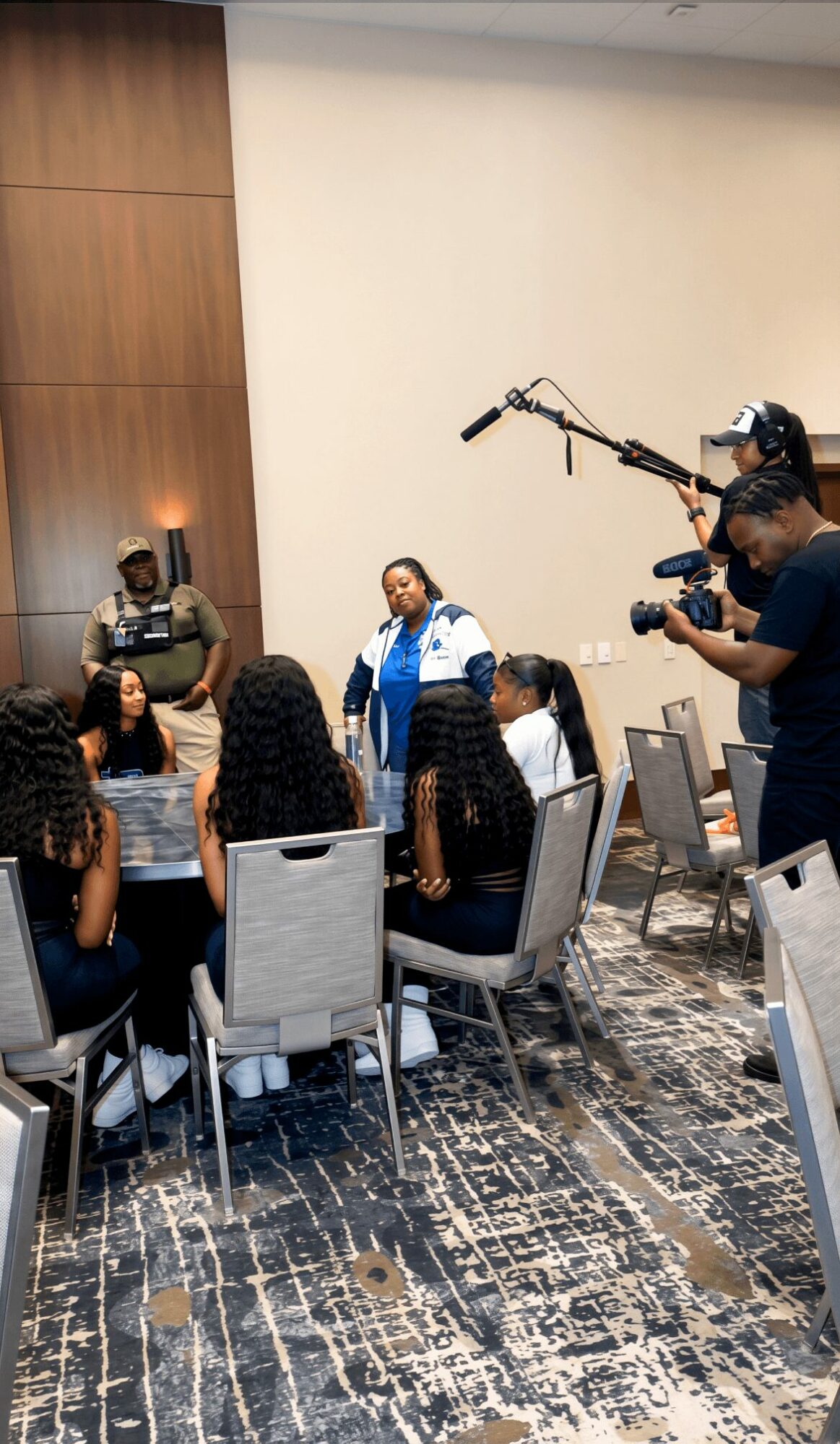 Group of women seated at a table in a conference room, with a woman speaking and crew filming her, camera operator nearby.