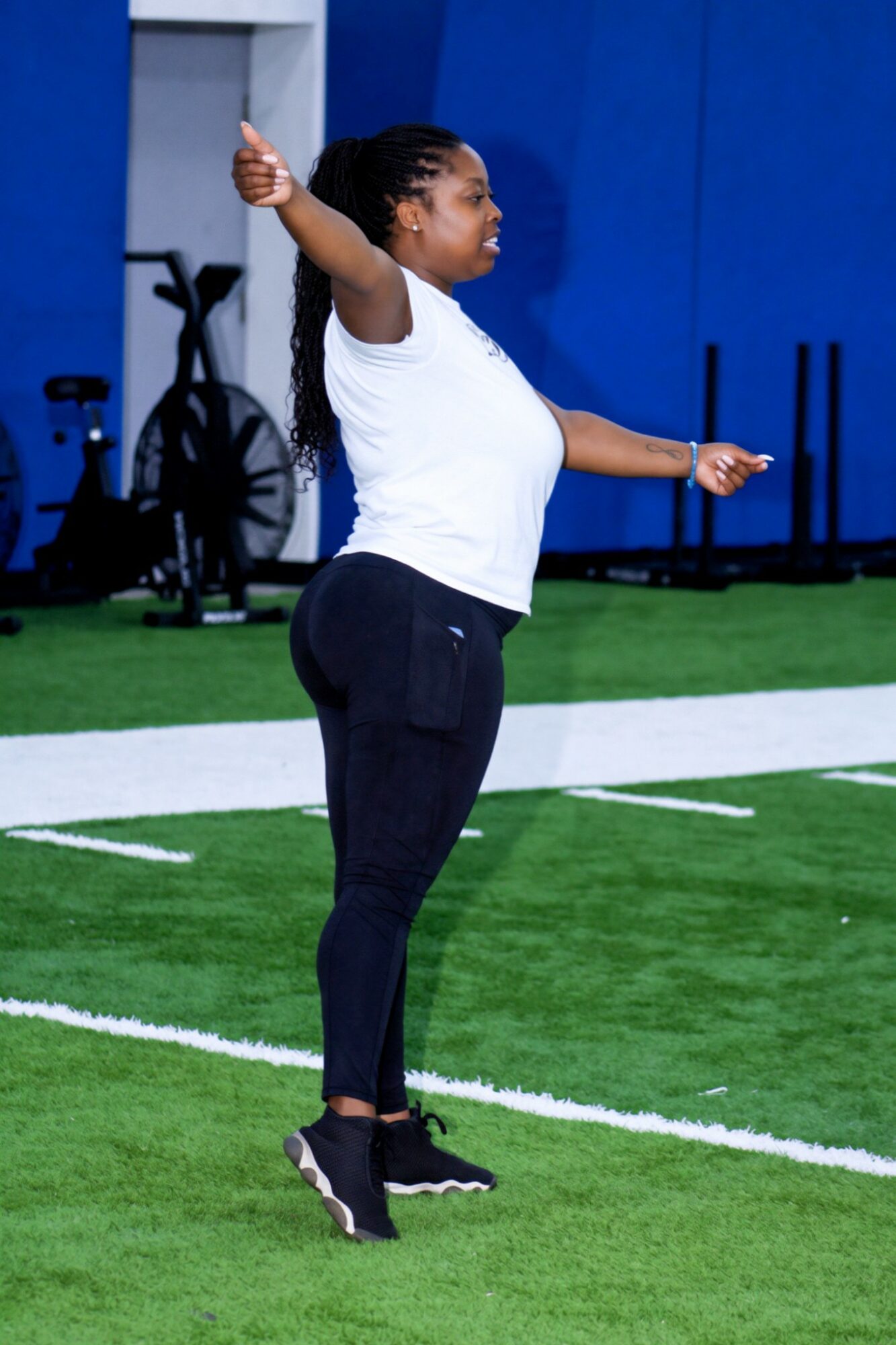 Woman standing on a sports field with arms raised, wearing a white shirt and black pants, in an indoor sports facility.