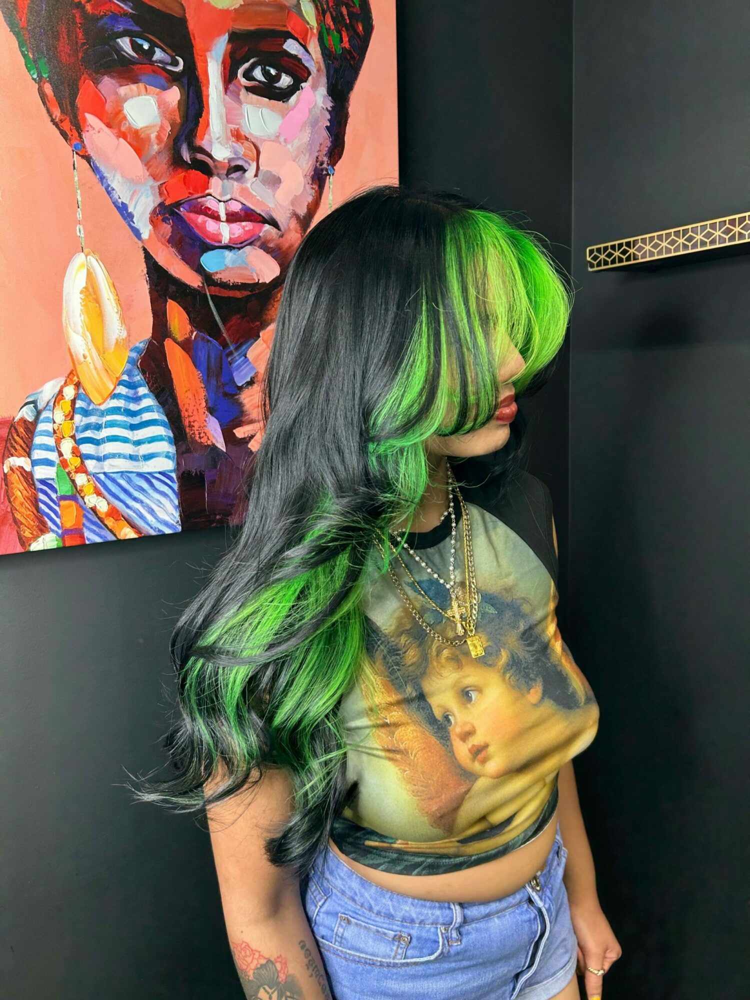 Person with long hair dyed green and black, wearing a graphic t-shirt and jeans, standing next to colorful portrait painting.