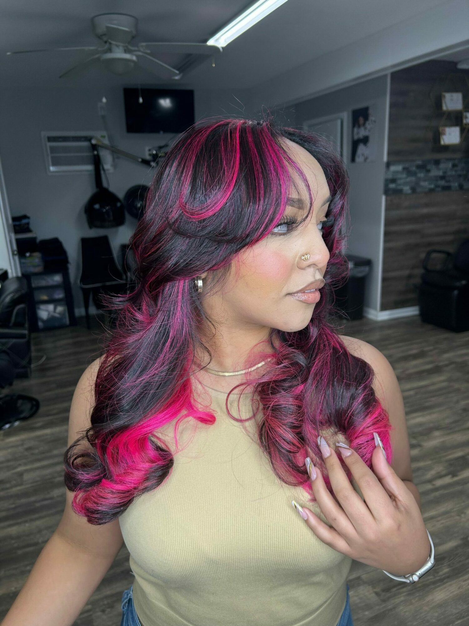 Woman with long, wavy hair dyed pink and black, wearing a beige top, in a room with wooden floor and shelves.