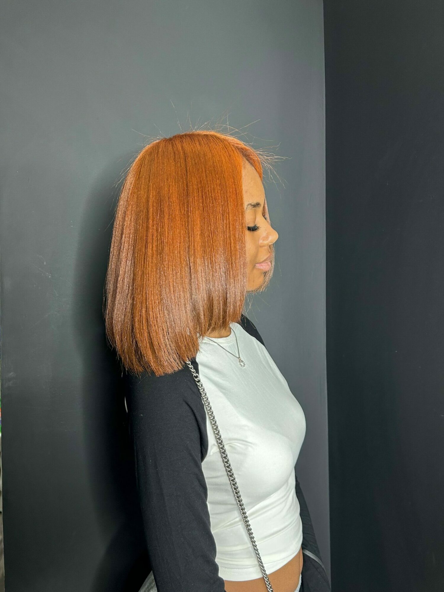Side view of a person with straight, shoulder-length orange hair, wearing a white top and black jacket, standing against a dark background.