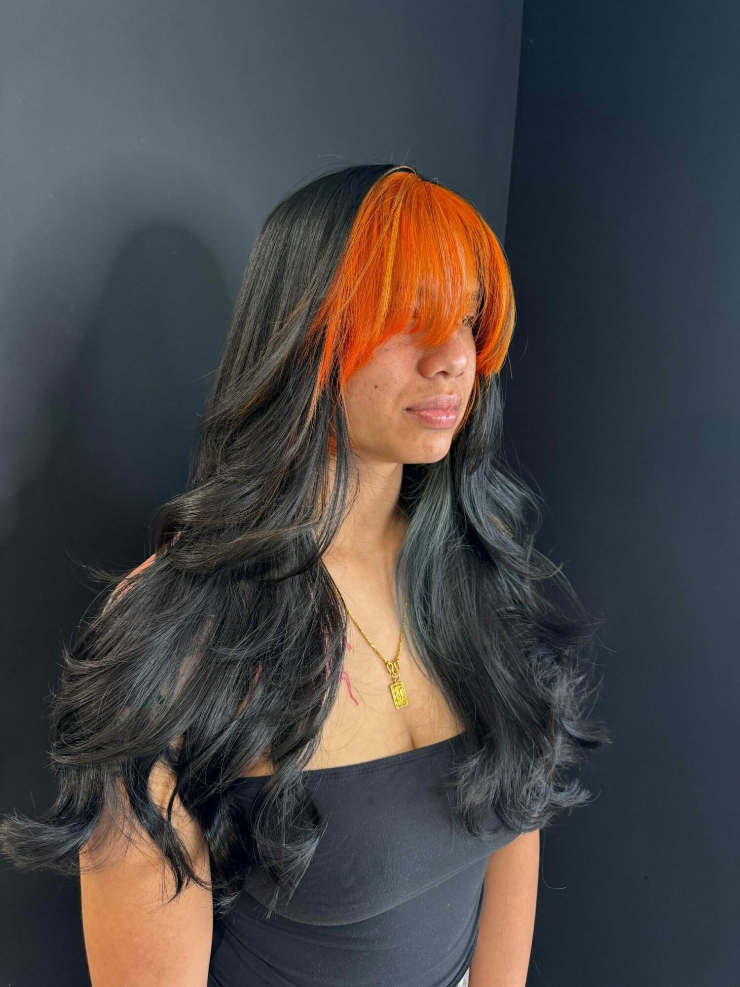 Woman with long black and orange hair lying on her side, wearing a black top, against a dark background.