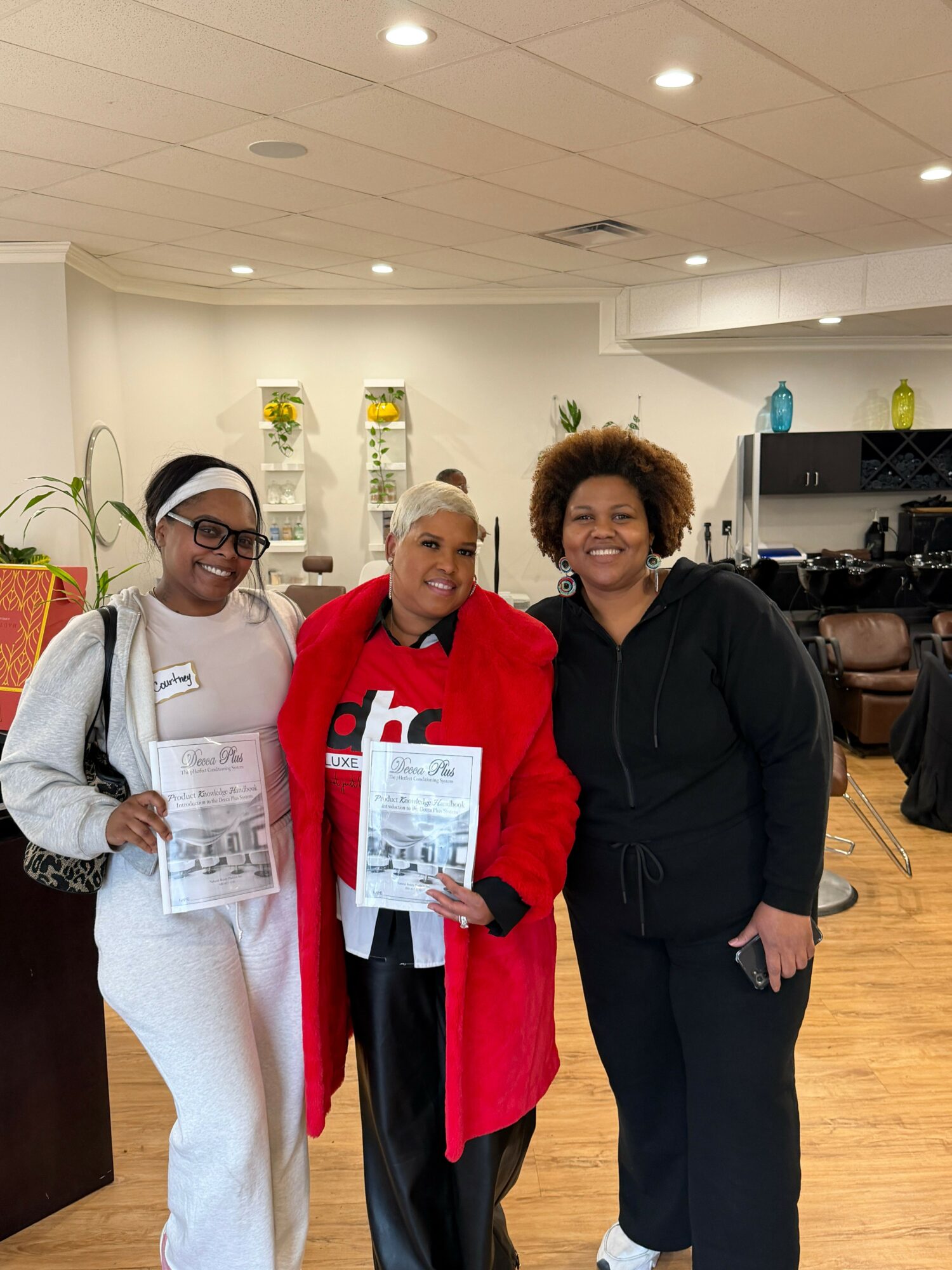 Two women and one girl standing indoors, one woman in black, girl in white, woman in red holding papers, smiling.