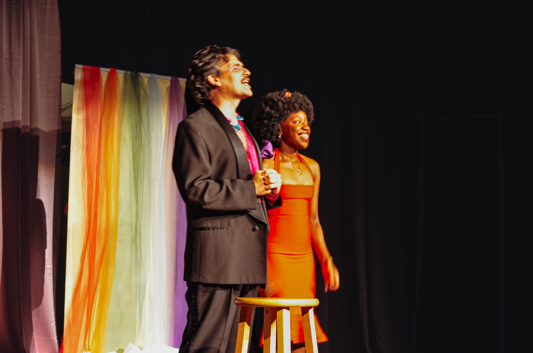 Two people stand on stage with a colorful curtain backdrop, one holding a microphone, the other smiling, a stool nearby.