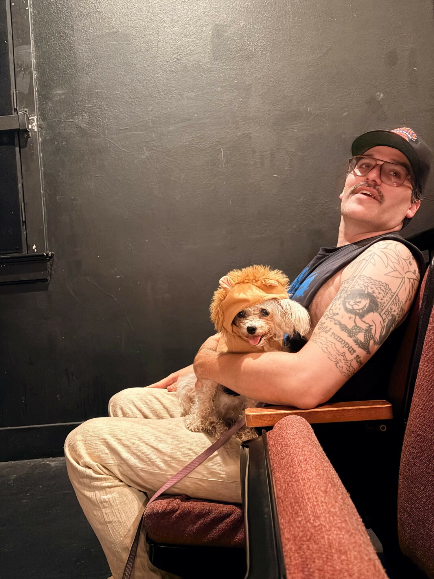 Person with glasses and tattoos holding a small dog, sitting on a chair in a room with a dark wall.