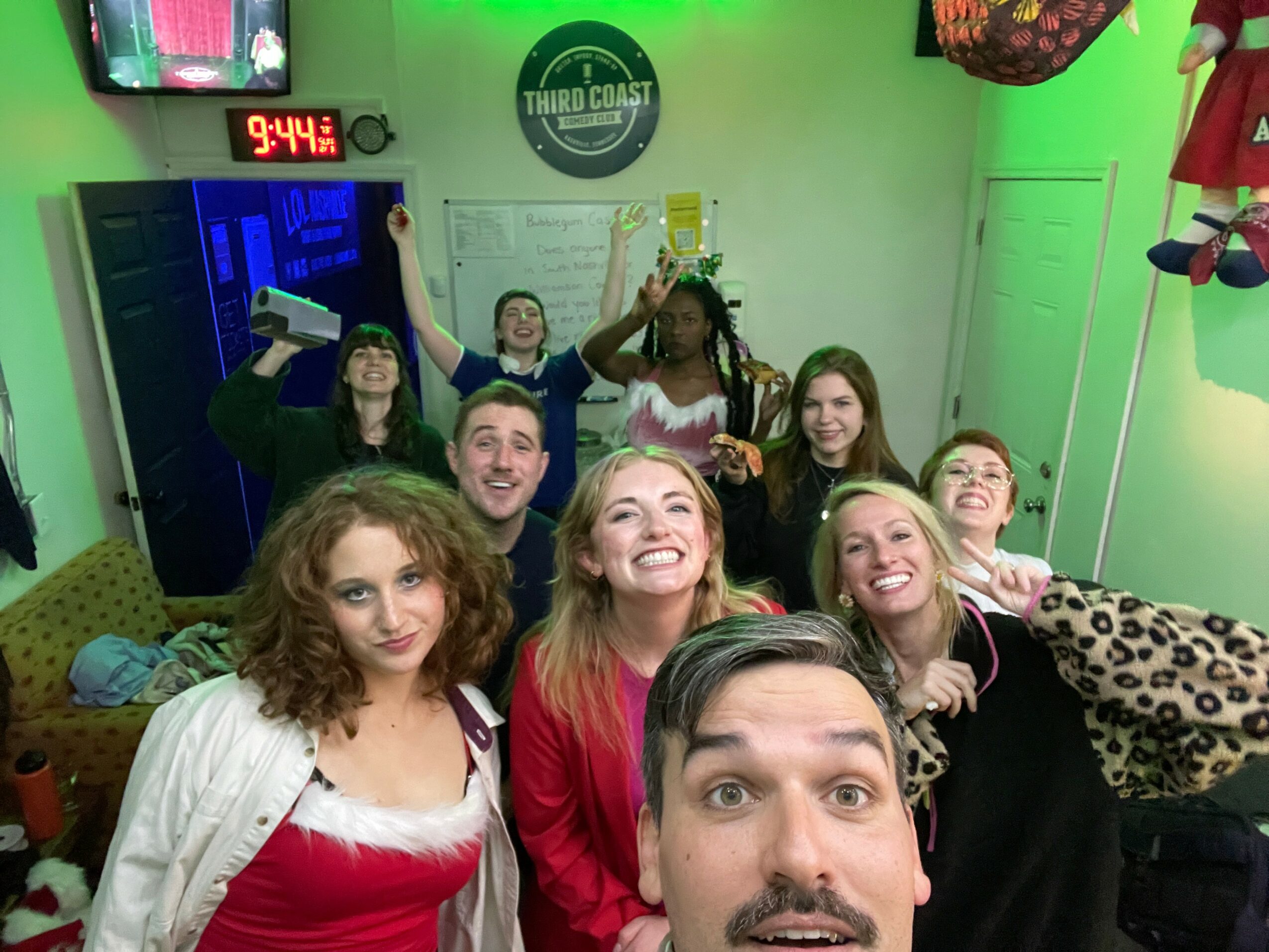 Group of nine people smiling and posing in a room with green walls, a TV, and a whiteboard, celebrating together.
