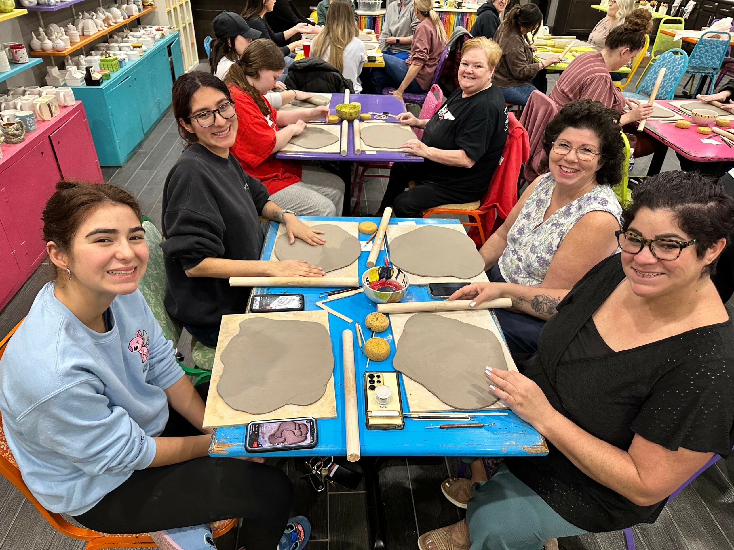 Group of women and girls sitting at tables in a colorful room, smiling, with craft supplies on the table.