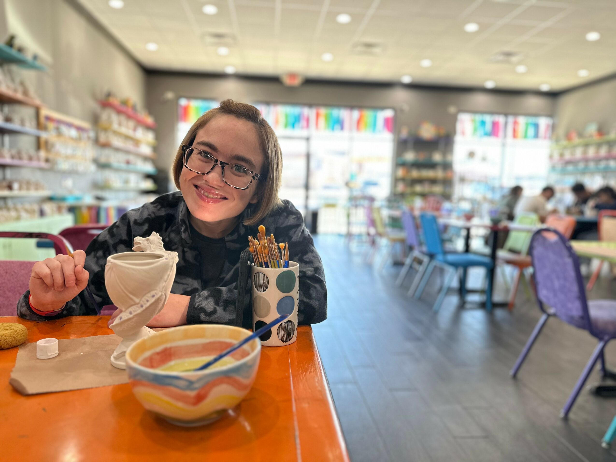 Smiling woman with glasses sitting at a table in a brightly lit store or cafe, with bowls and utensils on the table.