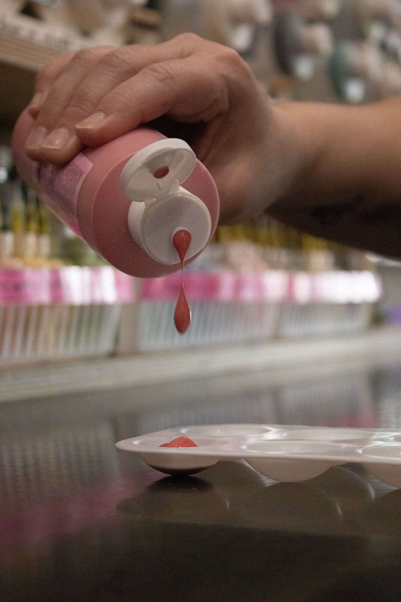Hand holding a pink bottle pouring a red liquid onto a plate, with shelves in the background.