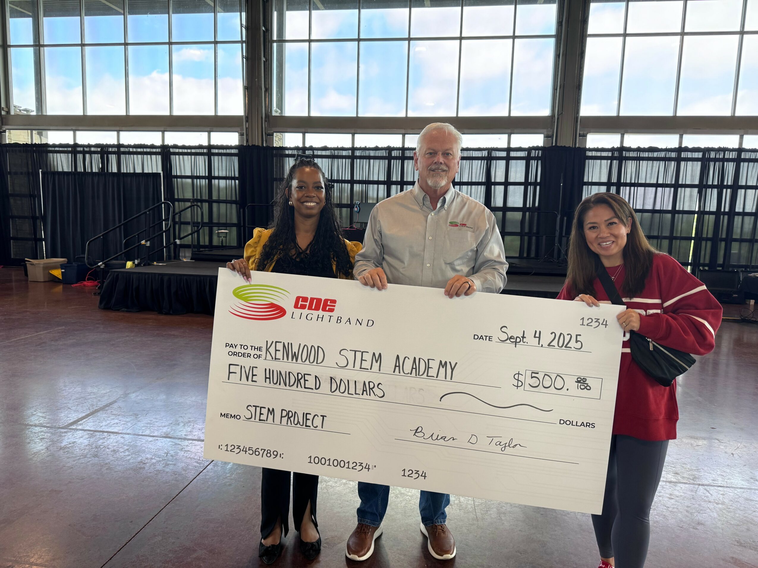 Three people holding a large check for $500 made out to Kenwood STEM Academy, dated September 4, 2025, in a spacious room with large windows.