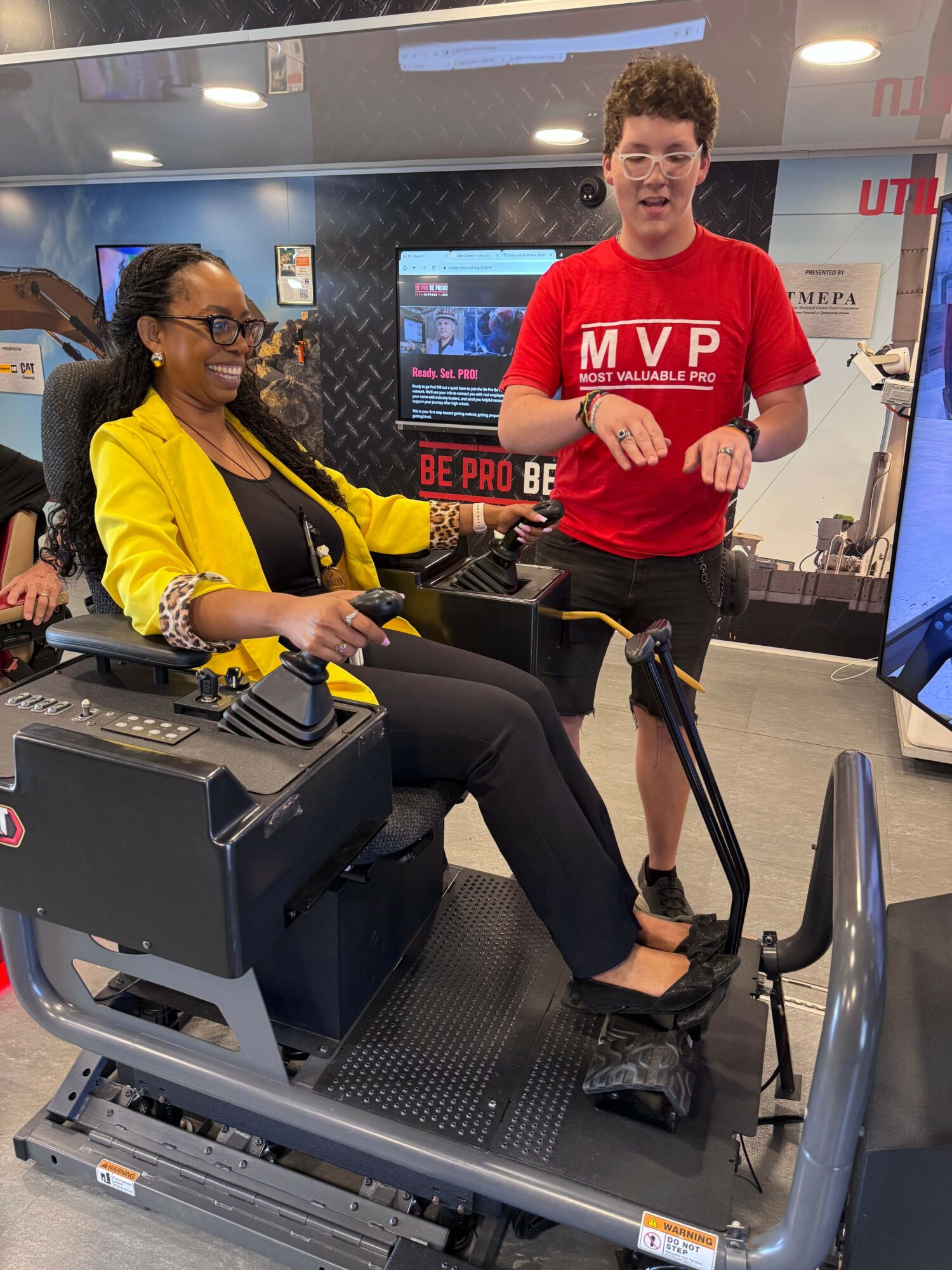 Woman using a treadmill in a gym, man standing nearby, gym equipment and screens in background.