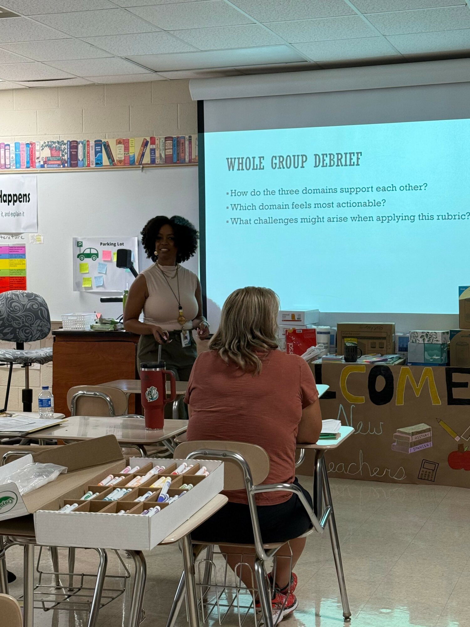 Two women stand in front of a large screen in a classroom or presentation room, with tables and chairs nearby.