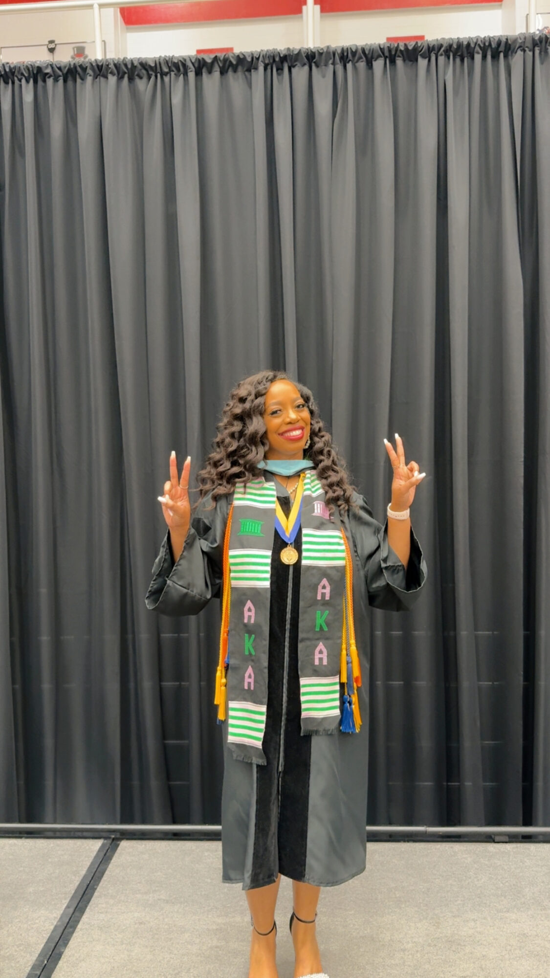 Person in graduation gown and stole, smiling and making peace signs, standing in front of black curtain backdrop.