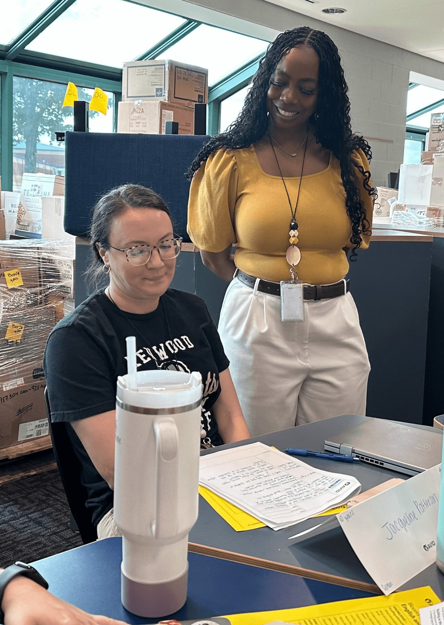 Two women in an office or store, one seated with glasses and a black shirt, the other standing in a yellow top and white pants, smiling.