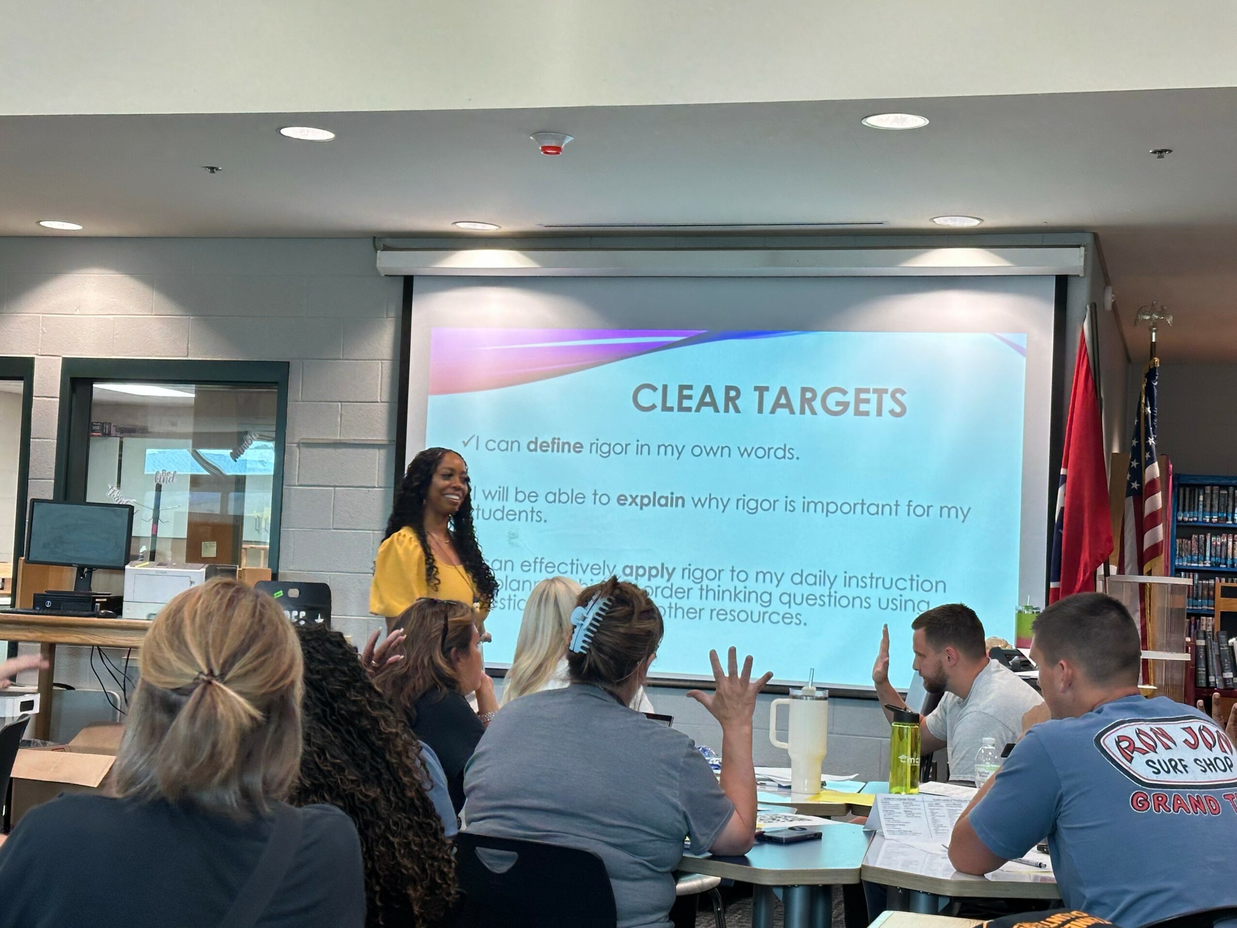 A woman presents in front of a projected slide titled 'Clear Targets' to a seated audience in a room with American flags.