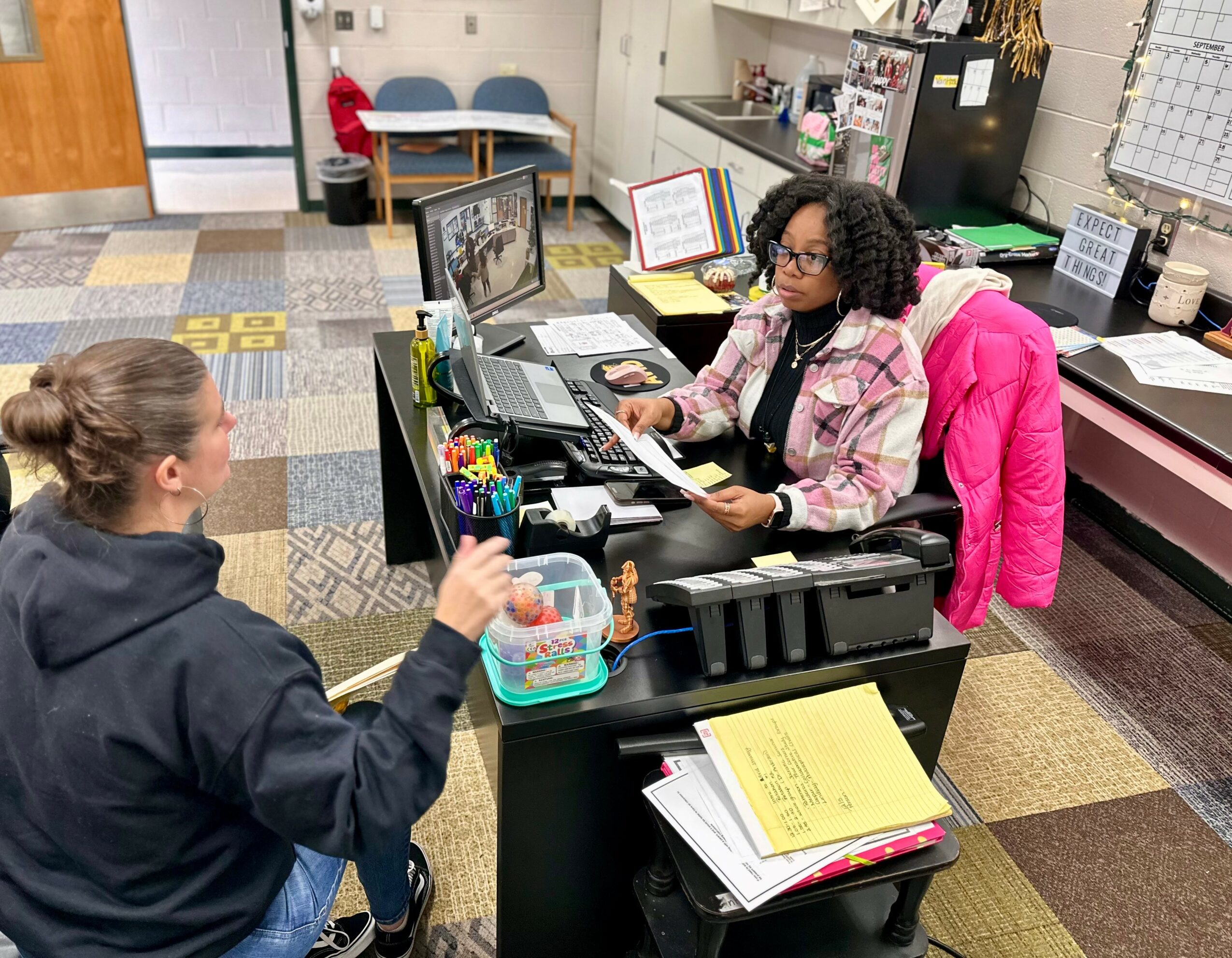 Two women in an office, one sitting at a desk with a pink jacket, the other standing with a black hoodie, engaging in conversation.