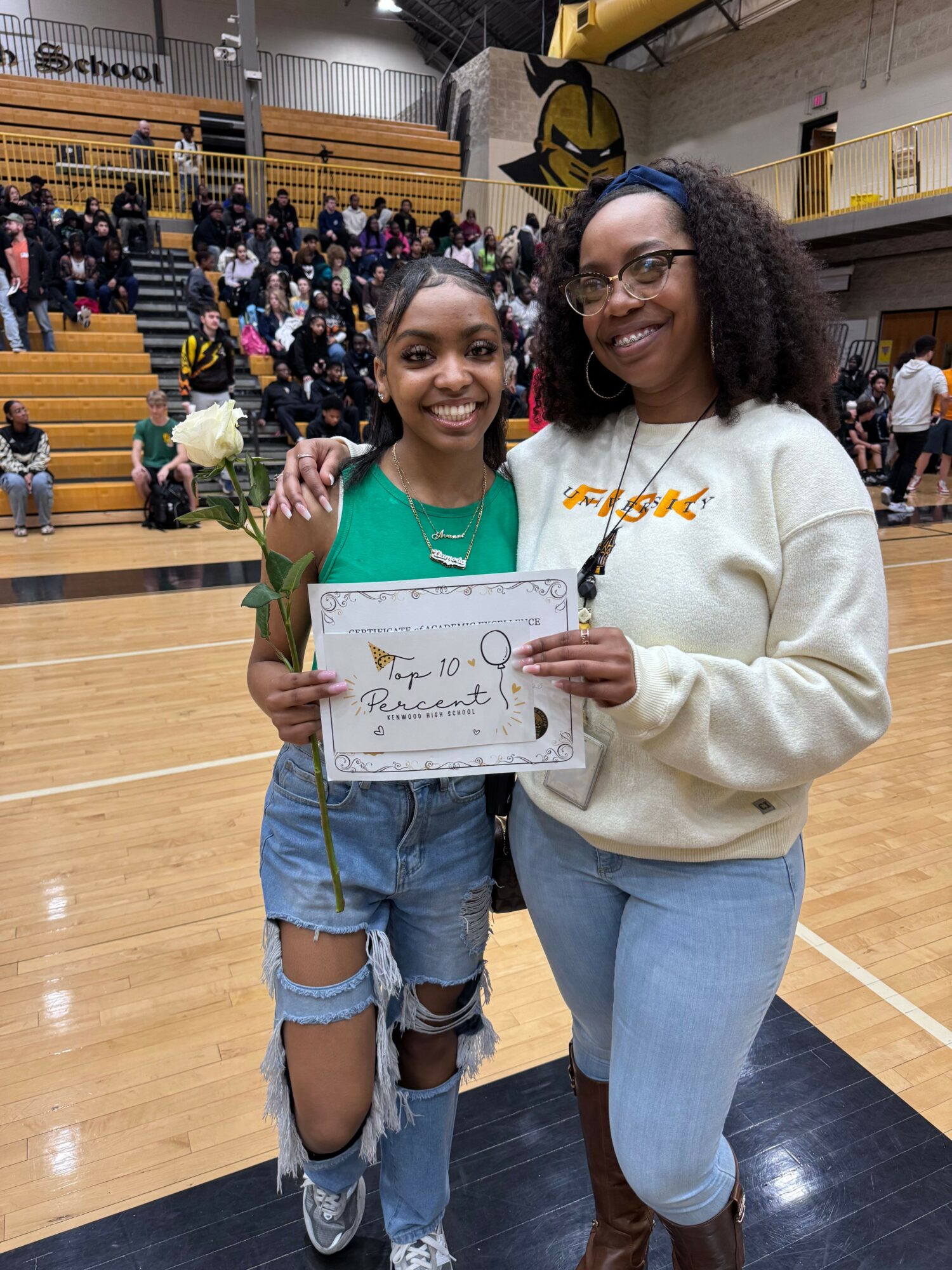 Two women standing indoors, one holding a certificate, with a crowd in the background. Both are smiling.