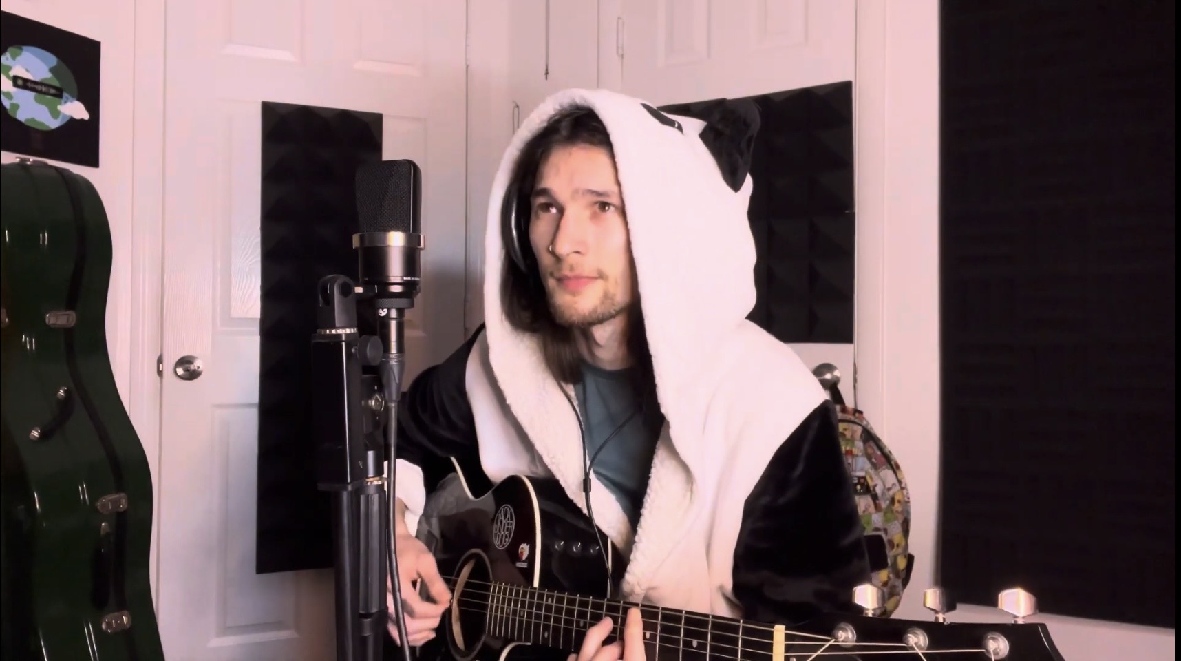 Person with dark hair wearing a hoodie with bear ears, holding a guitar in a music studio.