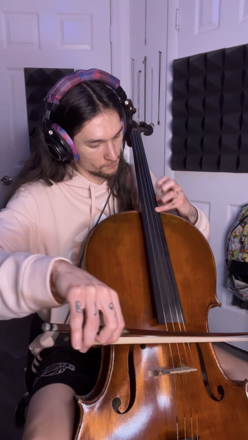 Person with long hair wearing headphones playing a cello in a room with soundproofing panels.