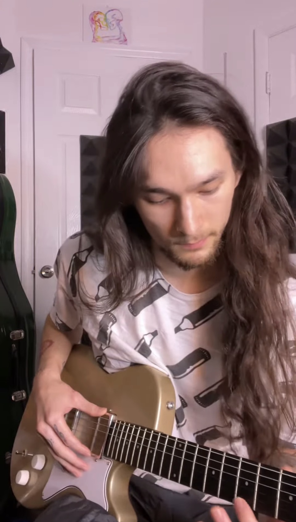 Person with long hair playing an electric guitar indoors, looking down at the instrument, with a pink wall and a drawing in the background.