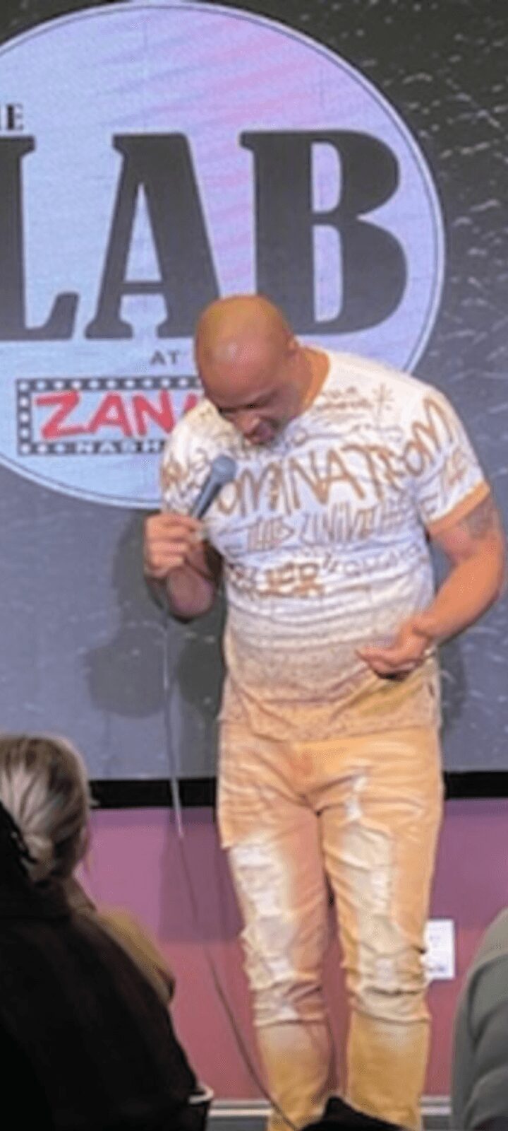 Bald man in a patterned shirt and light pants stands on stage, looking down, with a large circular sign behind him.