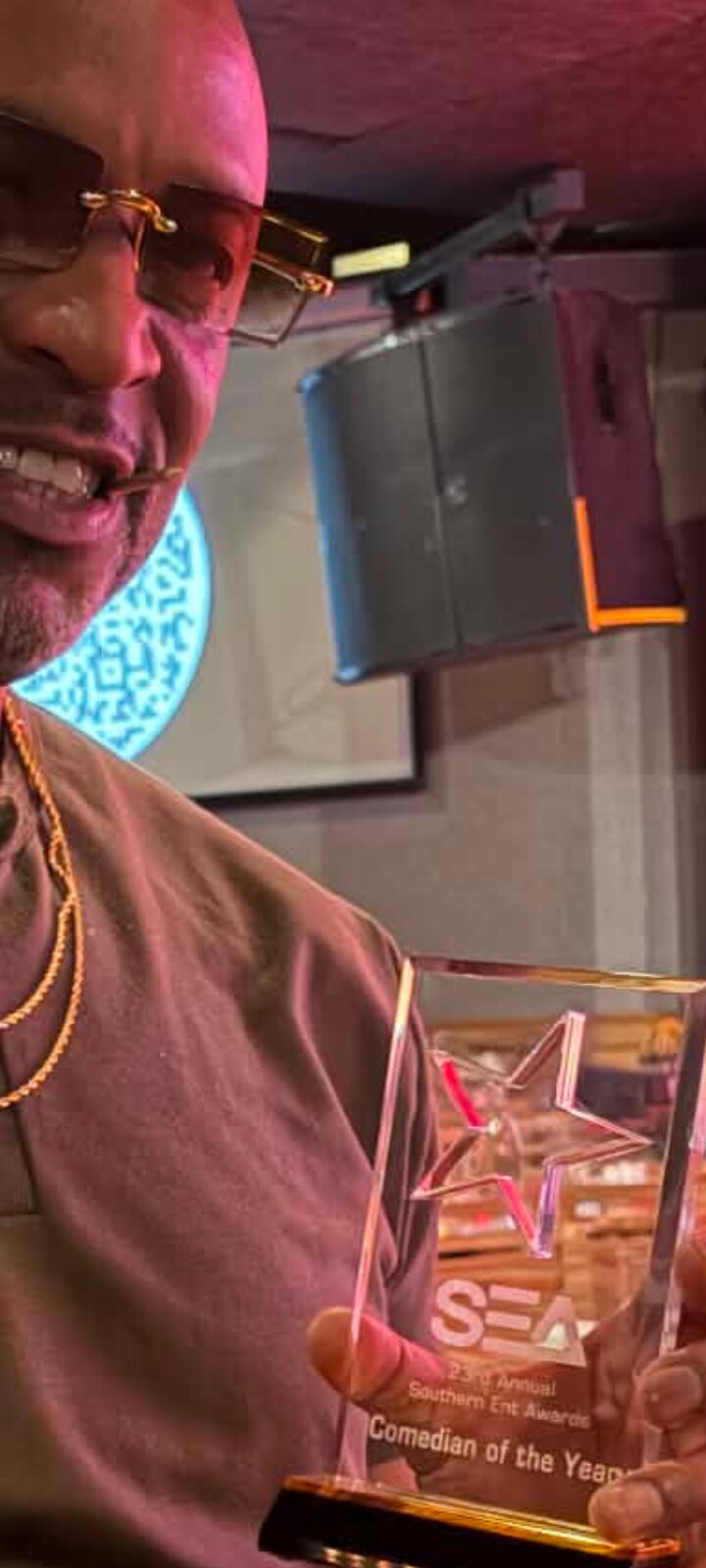 Close-up of a person wearing glasses and a gold chain, with a background of a speaker and a screen.