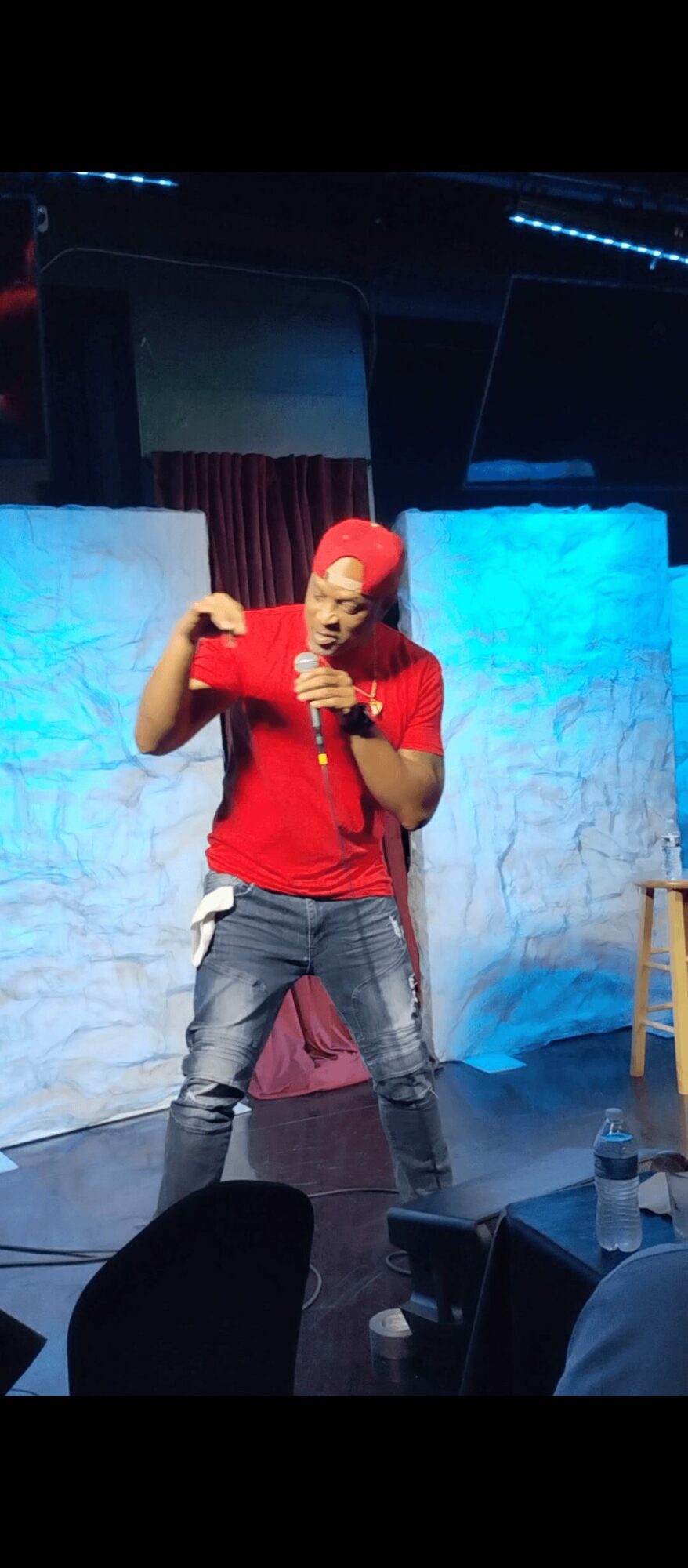 Person performing on stage with a microphone, wearing a red shirt and jeans, with blue and white background panels.