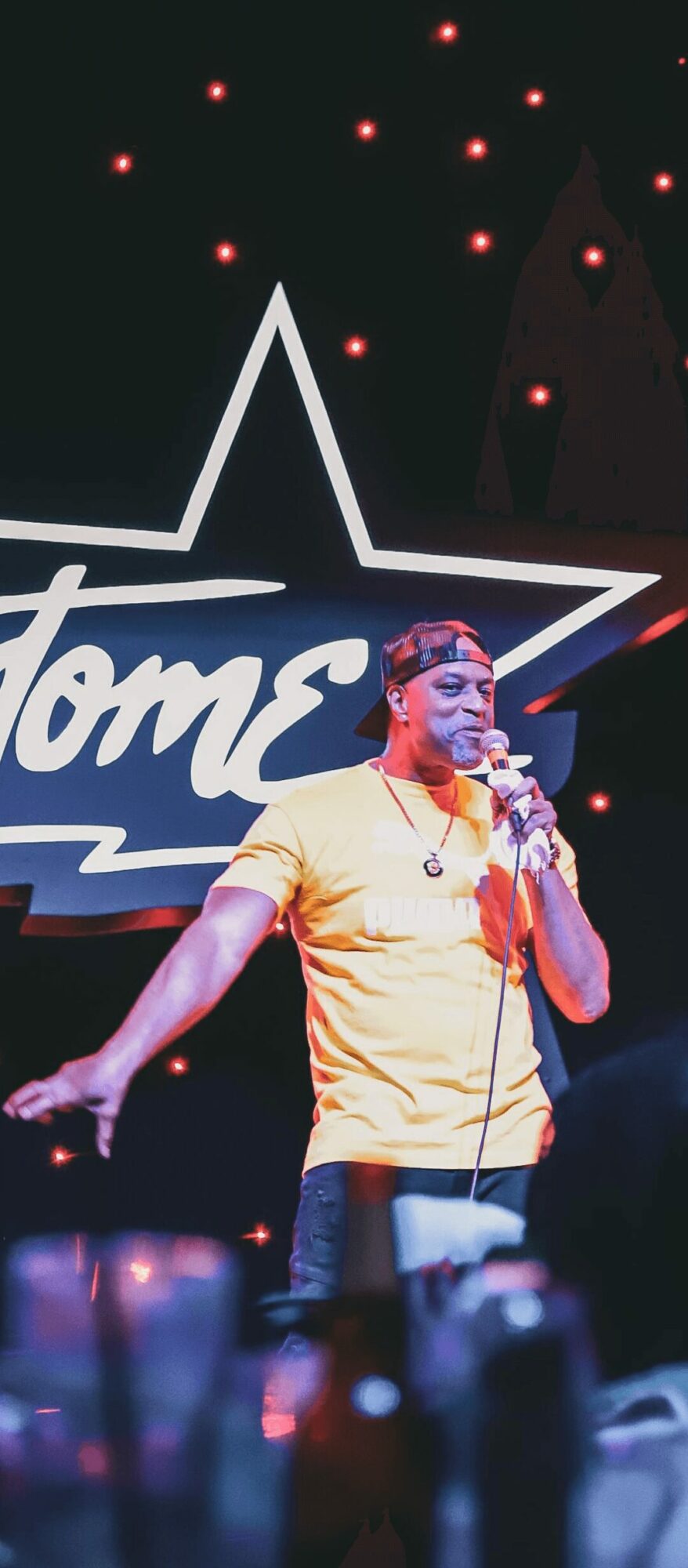 Performer on stage with a microphone, wearing a yellow shirt, in front of a star-shaped sign and a starry background.