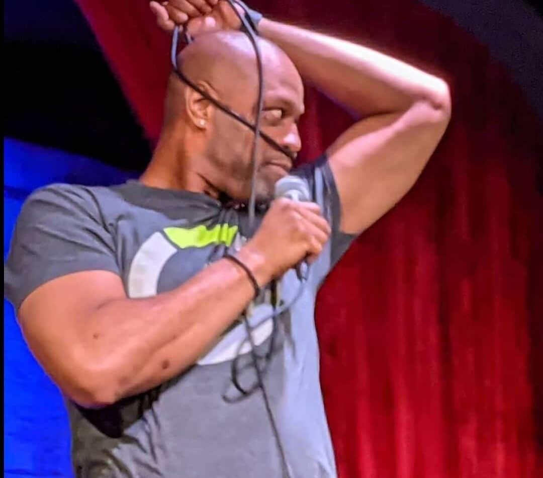 Person adjusting headphones with one arm raised, standing in front of a red curtain, wearing a gray T-shirt.